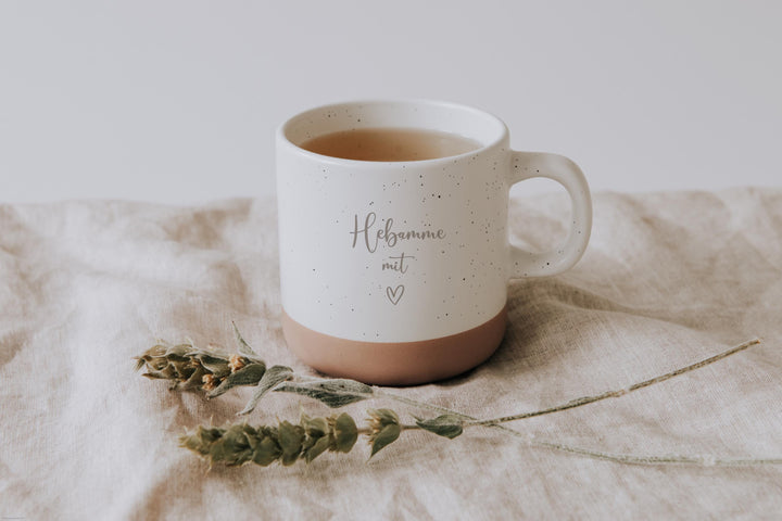 Steingut Tasse mit dickem Henkel als Geschenk und zum Danke sagen an die Hebammen.  Tasse mit weißer matter Lasur und kleinen sprenkeln der Oberfläche und unglasiertem Bodendesign in Natur, Graviert mit Hebamme mit Herz.